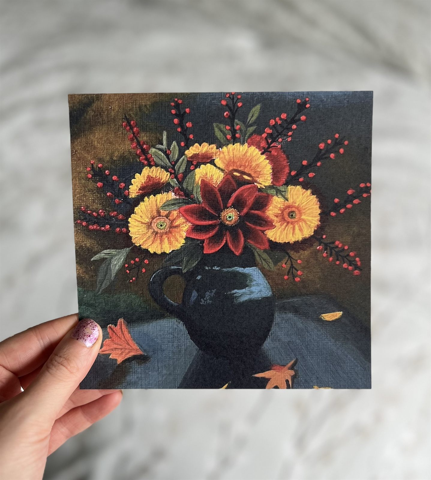 The Autumn Bloom print held in one hand: yellow and red autumn flowers with red berries in a dark green jug, with fallen leaves on the table