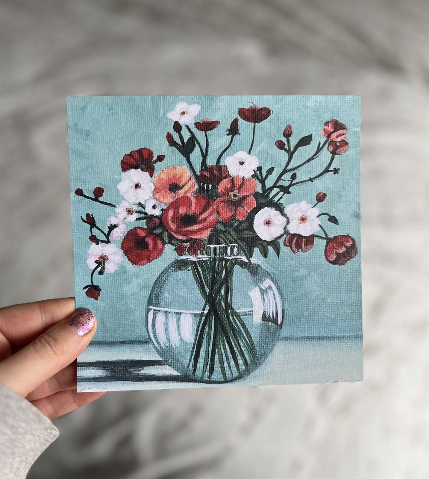 The Gathered Blooms print held in one hand: red poppies, peach blooms and white blossom in a round glass vase against a pale green background