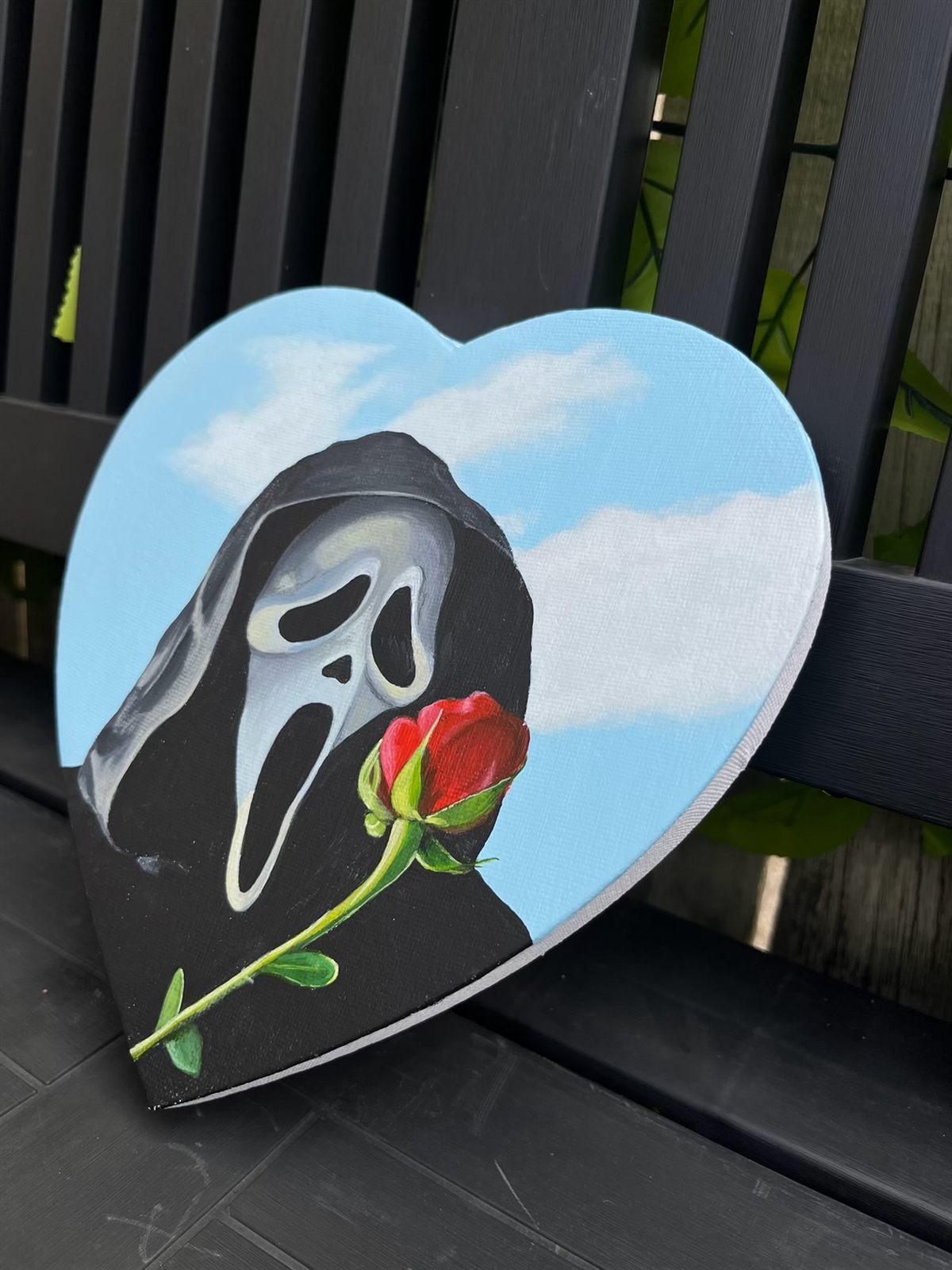 Ghostface, an acrylic painting on a heart-shaped canvas: the Ghostface mask in its black hood against a blue sky with white clouds, holding a long-stemmed red rose