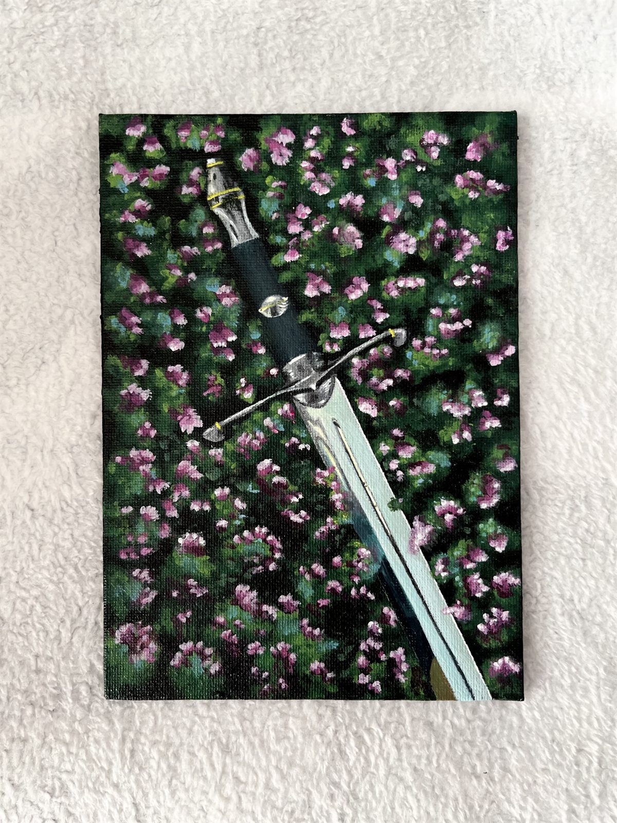 Lost Sword, an acrylic painting on a small canvas panel: a sword lying point-down across a dark field scattered with pink and white flowers
