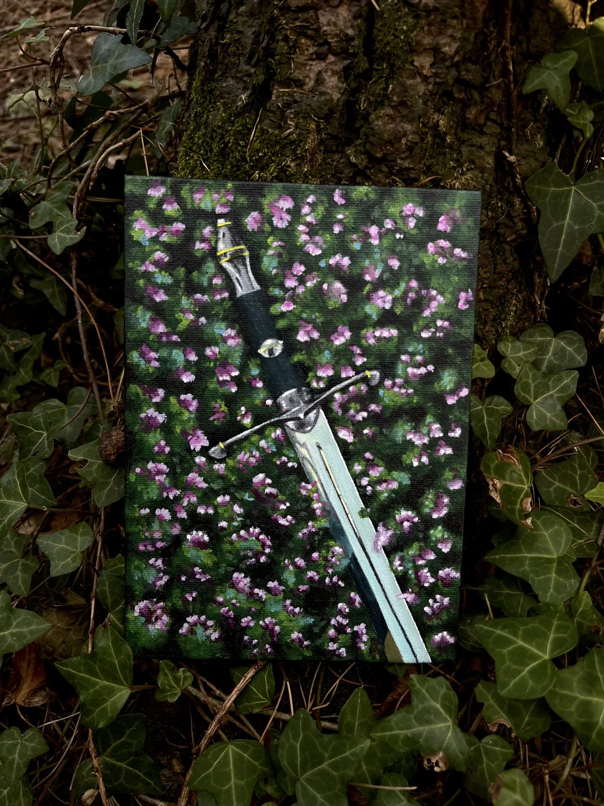 An acrylic painting on a small canvas panel: a sword lying point-down across a dark field scattered with pink and white flowers
