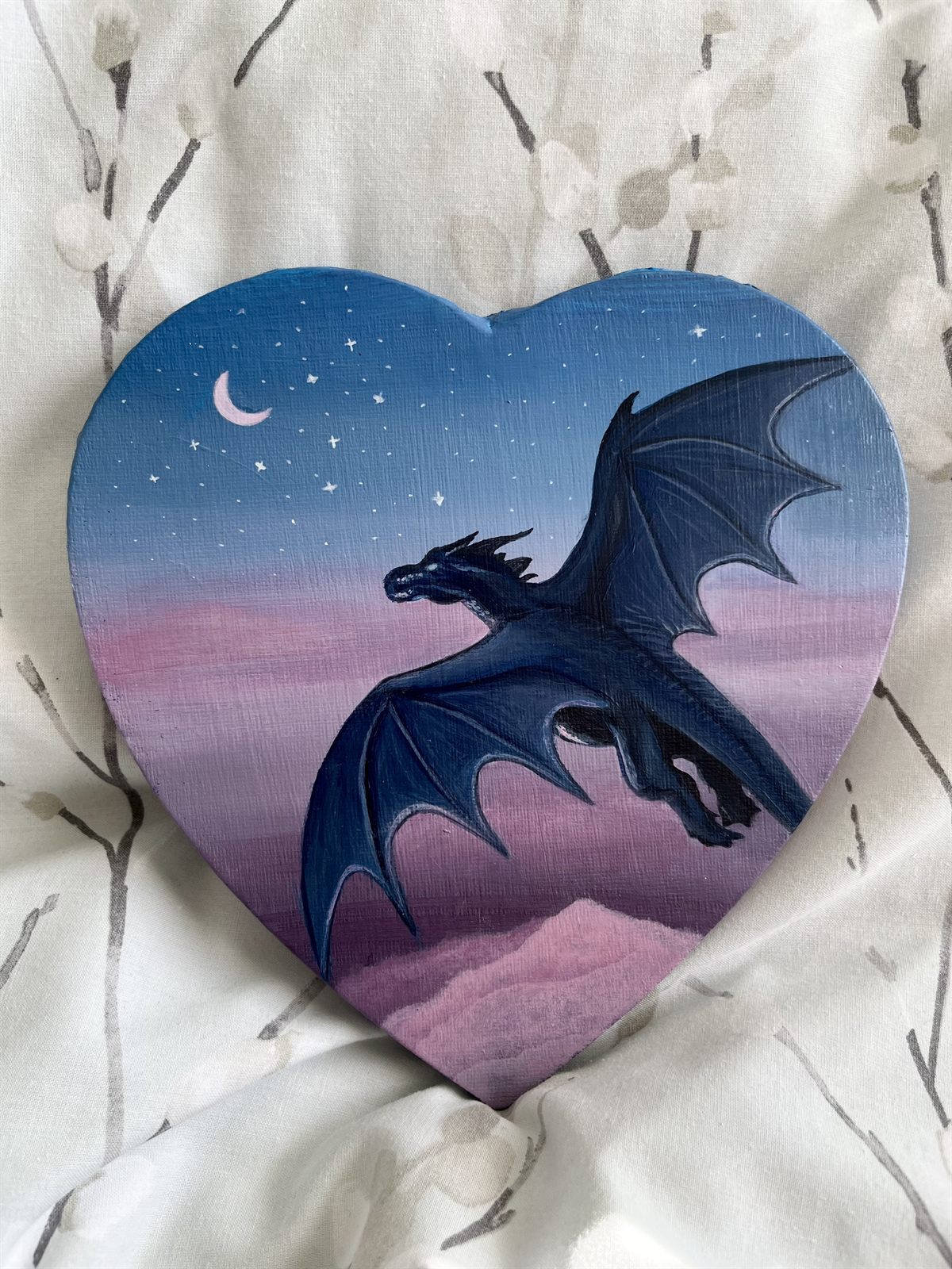 Nightwing, an acrylic painting on a heart-shaped canvas: a dark blue dragon in flight over pink clouds, against a starry dusk sky with a pink crescent moon