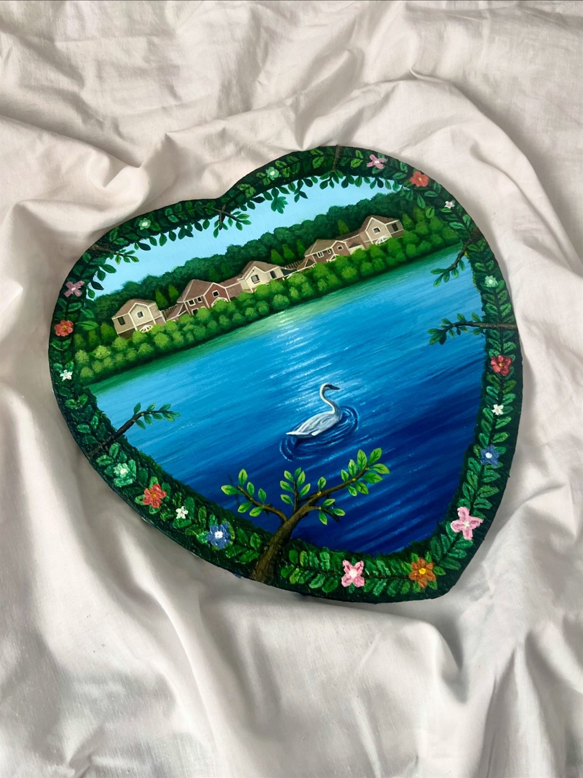 Peace, an acrylic painting on a heart-shaped canvas: a swan on a blue lake with houses and trees along the far bank, framed by a painted border of leaves and flowers