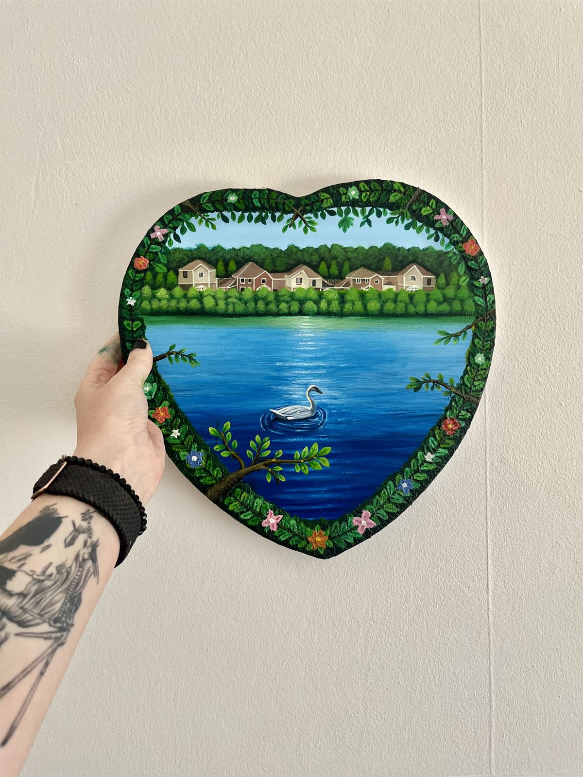 Peace, an acrylic painting on a heart-shaped canvas: a swan on a lake with houses and trees on the far bank, framed by a painted border of leaves and flowers