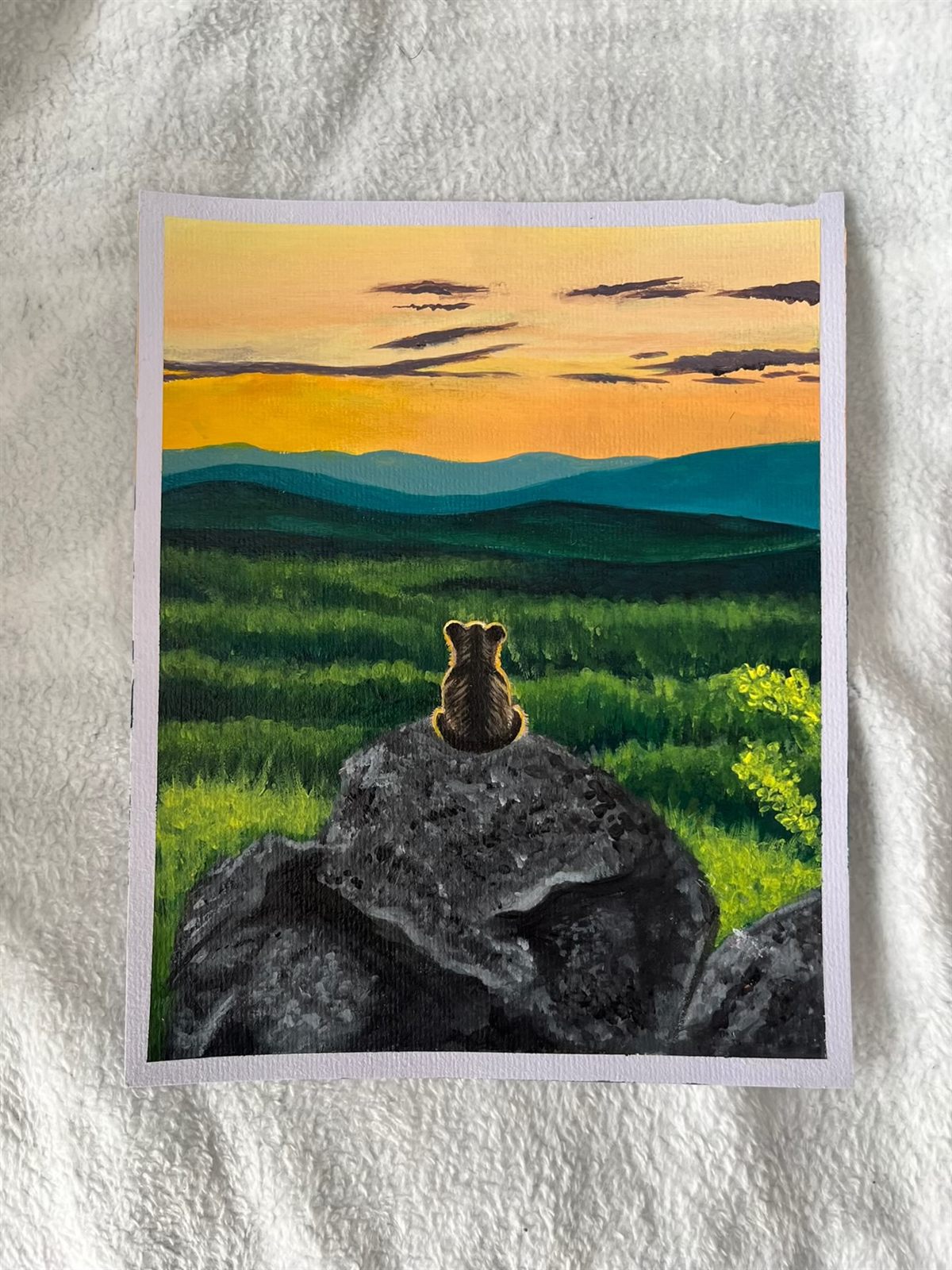 Rest Stop, an acrylic painting of a bear cub sitting on a rock with its back to you, looking out over green hills towards a golden sunset